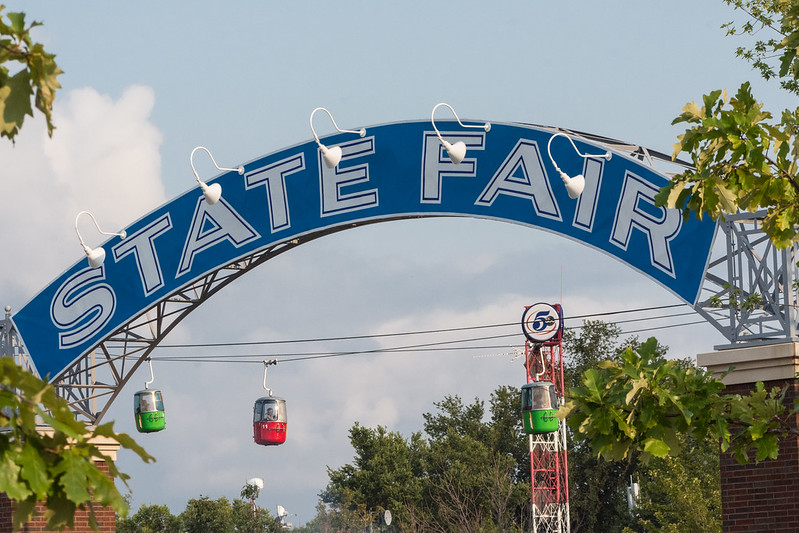 Banner over entrance to Minnesota State Fair reads "State Fair," elevated passenger tram cars in background
