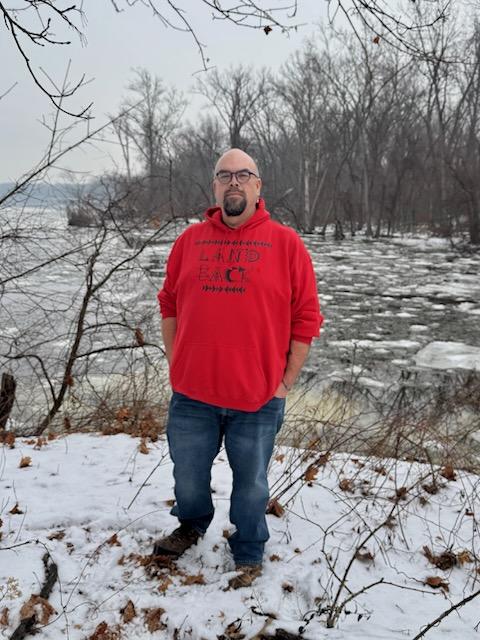 Indigenous man in red #Landback hoodie stands in a wintry, lightly forested landscape