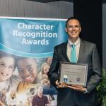 A dashing man holding a plaque stands next to a pull-up graphic stand that reads "Character Recognition Awards"