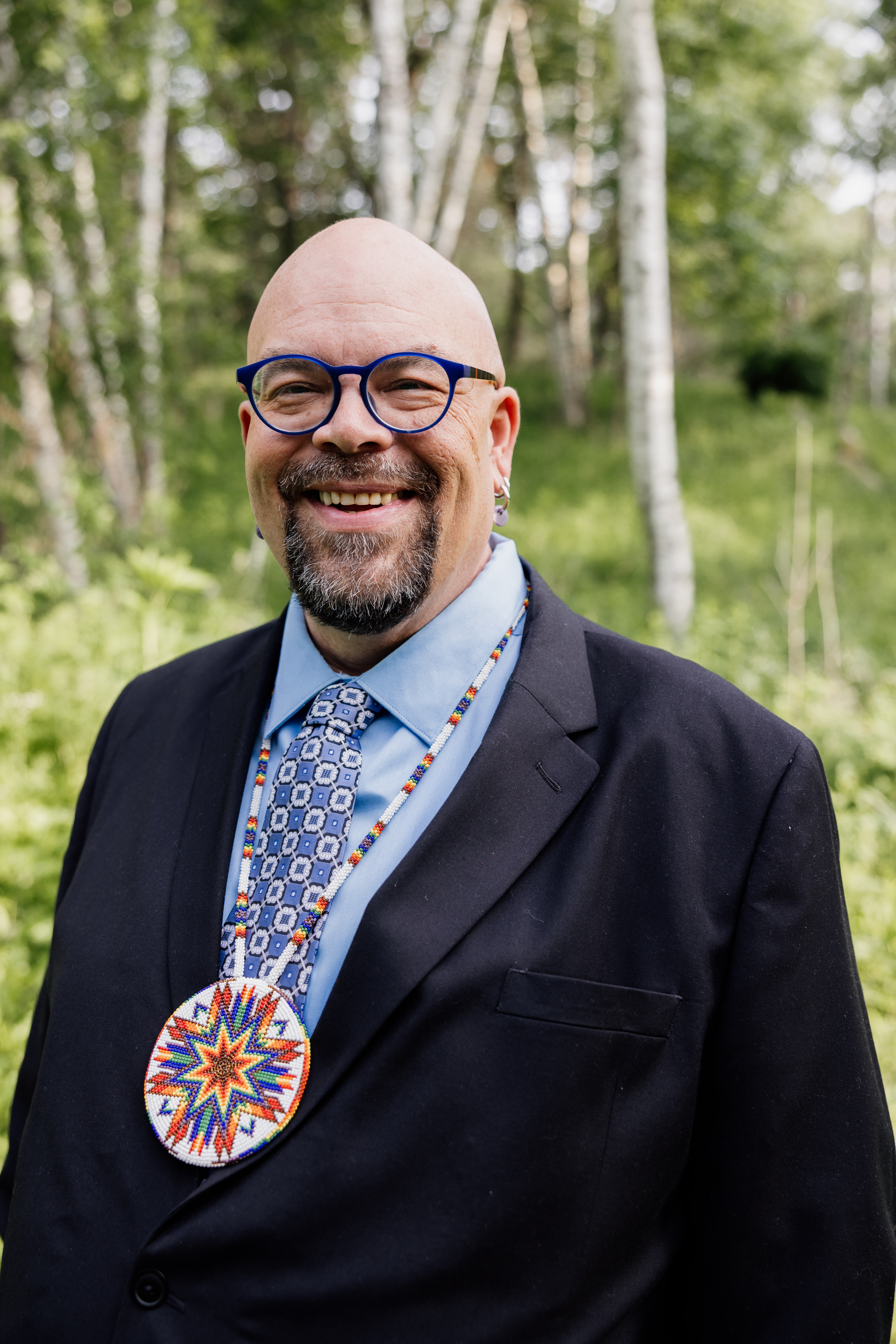 A man in a suit with an indigenour art necklace stands, smiling and looking at the camera, in an outdoor forested setting