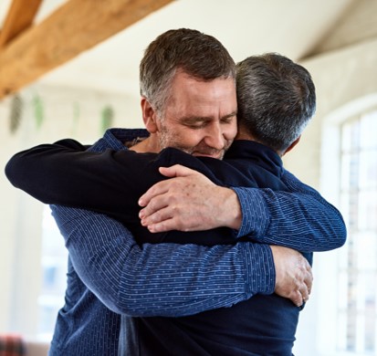 Two middle-aged men hug in a sunlit house.
