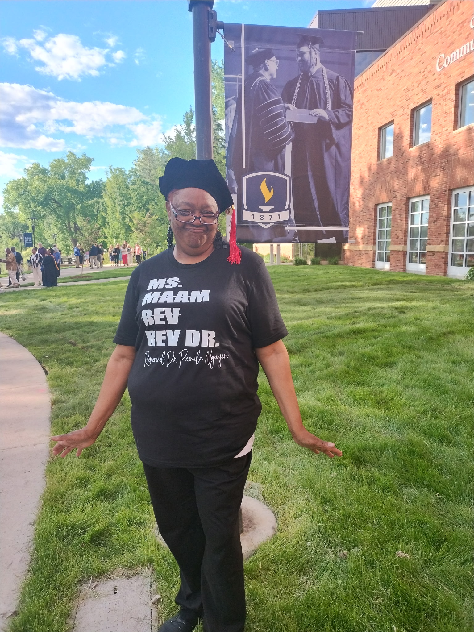 A newly-minted graduate stands outdoors, posing in her graduation cap and a black t-shirt that reads, in four stacked rows of white text, "Ms. / Maam / Rev. / Rev. Dr." The first three rows are struck through. Below the last row, in cursive, is her name