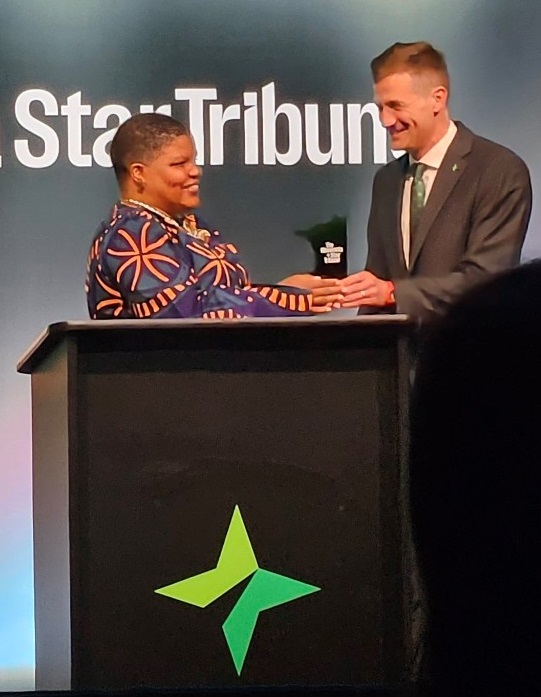 A woman with close-cropped hair and a beautiful purple dress with orange lines receives an award at a podium from a man in a suit and tie. The word "Star Tribune" is on the wall behind them. 