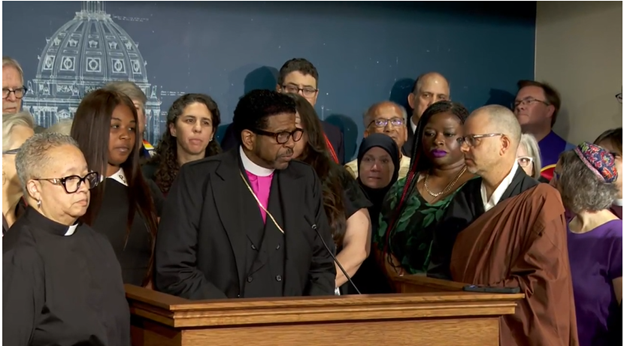 A man in a black suit with purple shirt and white collar stands at a podium, flanked by dozens of other people in religious garb.