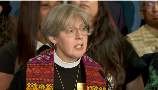 A woman in glasses wearing a black robe, white collar, and orange and yellow stole speaks at a podium in a close-up image