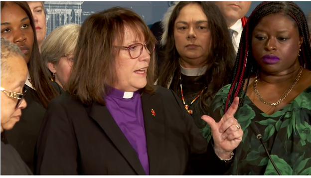 A woman in a black jacket, purple shirt and white collar, lifting her left hand in a gesture, speaks at a podium