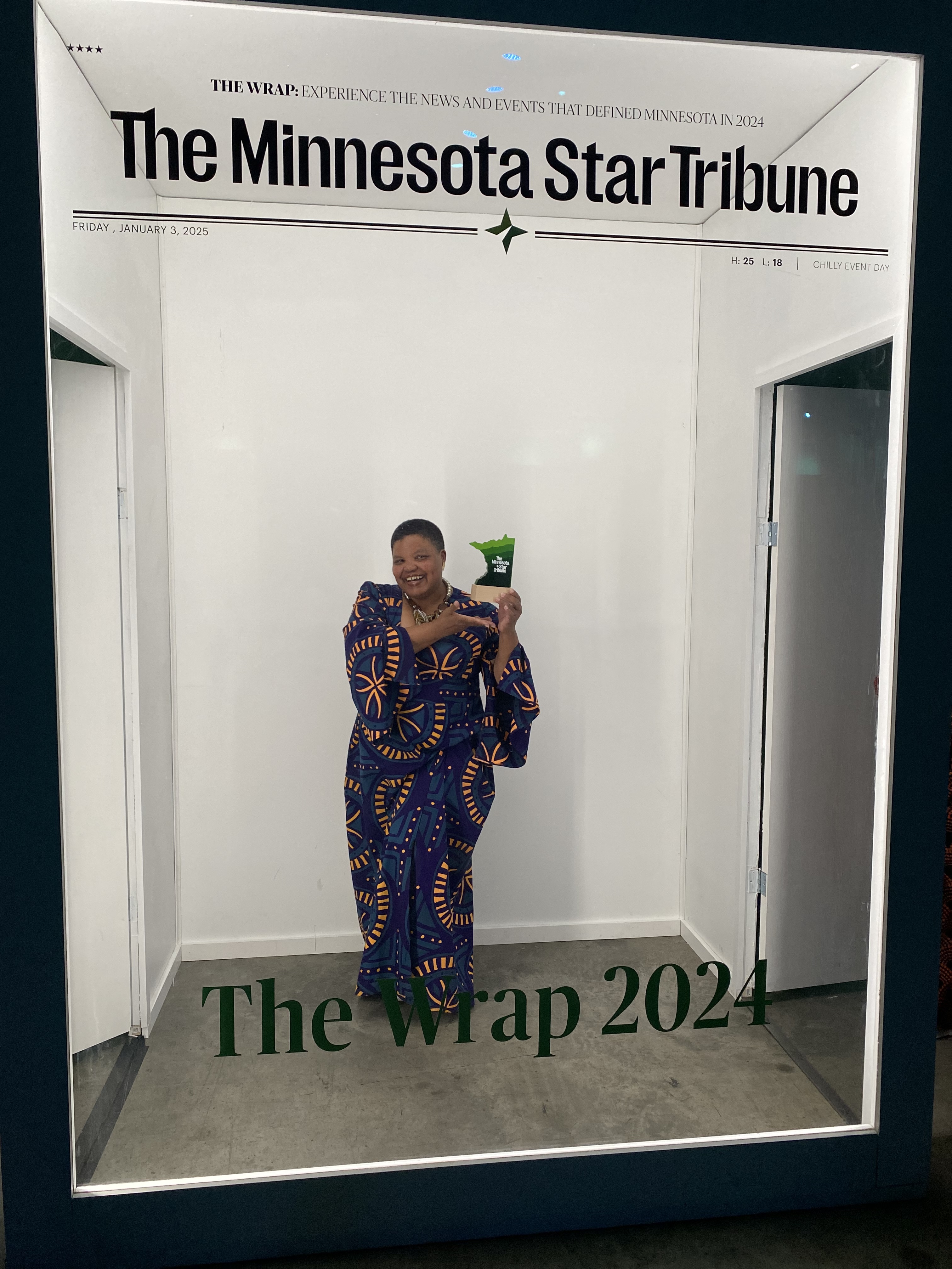 A woman in a beautiful purple dress with orange lines jauntily holds an award behind a glass wall made to look as though the person behind it is on the cover of the Minnesota Star Tribune.