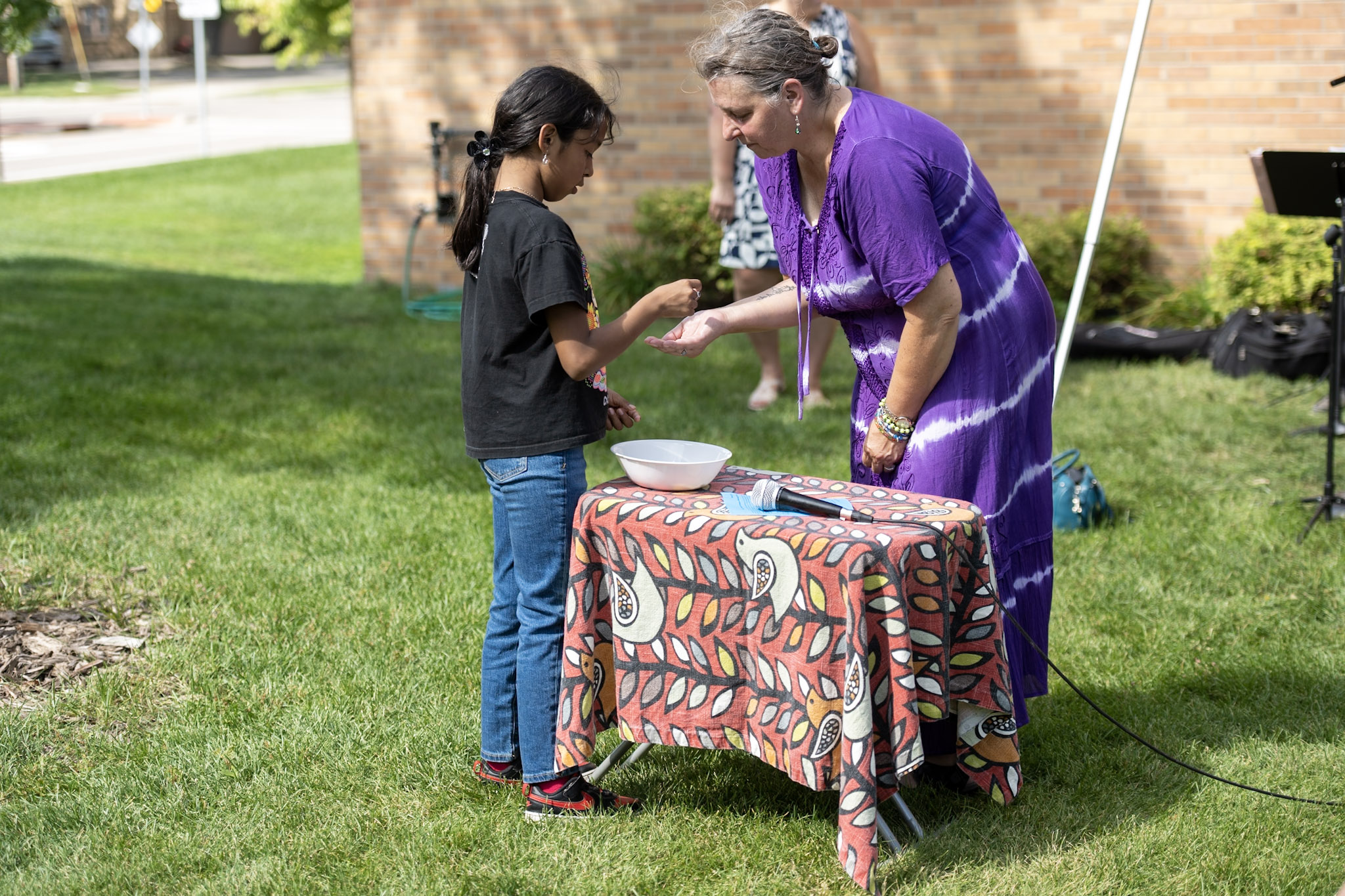 A woman in a patterned purple dress bends slightly to receive communion bread across a table from a girl in a black shirt. Both are outisde.