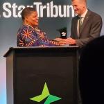 A woman with close-cropped hair and a beautiful purple dress with orange lines receives an award at a podium from a man in a suit and tie. The word "Star Tribune" is on the wall behind them. 