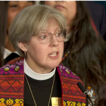 A woman in glasses wearing a black robe, white collar, and orange and yellow stole speaks at a podium in a close-up image