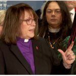 A woman in a black jacket, purple shirt and white collar, lifting her left hand in a gesture, speaks at a podium