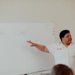 A woman points to a whiteboard while facing an off-screen audience in a brightly-lit room